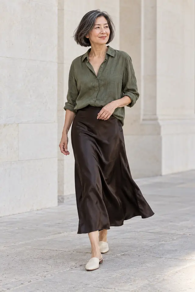 Mujer de cabello oscuro con canas, blusa oliva, falda midi de satén ébano y zapatos planos marfil.