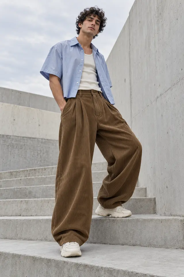 Hombre con pantalón de pana tabaco de gran volumen, camisa corta azul claro abierta sobre una camiseta de tirantes marfil y zapatillas claras.