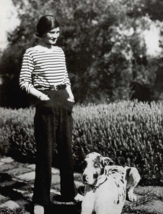 Gabrielle Coco Chanel con una camiseta marinera y pantalón oscuro, acompañada por un perro, hacia 1928.
