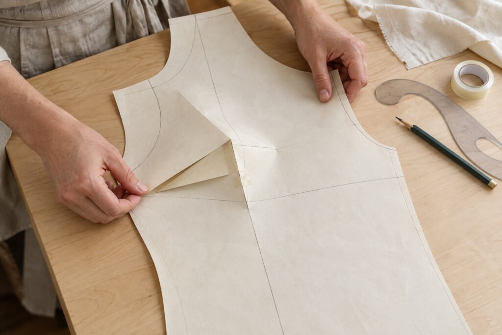 Manos manipulando una pieza de patrón con papel plegado y cinta adhesiva, junto a un lápiz y una regla curva sobre la mesa.